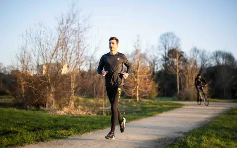 Un jeune homme s'entraîne à la course à pied sur un chemin ensoleillé, entouré d'arbres dénudés. Un cycliste passe en arrière-plan.