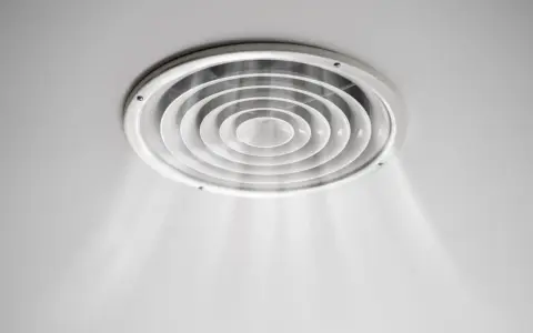 Grille de ventilation ronde au plafond, diffusant une légère brume. On dirait que la VMC pas assez puissante peine à renouveler l'air.