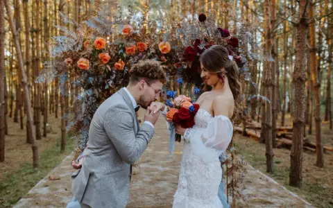Romantyczny ślub w lesie. Pan całuje dłoń panny młodej, która trzyma bukiet czerwonych i pomarańczowych róż.