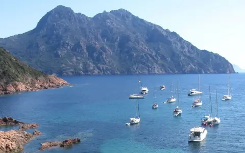 Vue d'une baie rocheuse avec plusieurs voiliers et bateaux à moteur en mouillage forain, sous une montagne imposante.