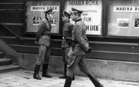 Trzej żołnierze w mundurach stoją przed kinem z plakatem Mariki Rökk. Stawka większa niż życie, teatr historii.
