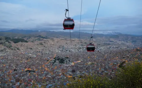 Red la paz gondola cabins glide over a sprawling city at dusk, with lights twinkling below and mountains in the distance.