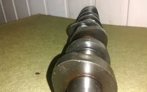Metalowy, wytarty wałek rozrządu leży na zielonym materiale. Widać ślady zużycia na jego krzywkach.