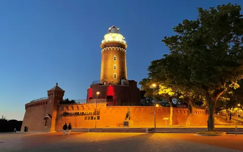 Wieczorny spacer przy latarni morskiej Kołobrzeg. Oświetlona ceglana budowla z czerwonymi akcentami i drzewa tworzą magiczny klimat.