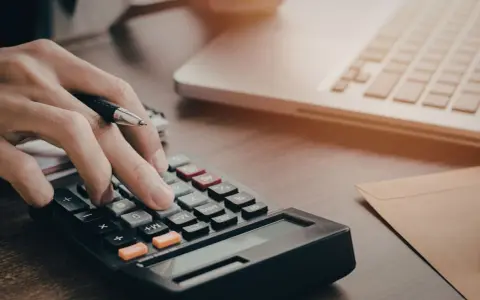 Osoba liczy na kalkulatorze, zastanawiając się, czy studia stacjonarne są płatne. Obok leży laptop i koperta.
