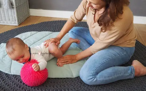 Fizjoterapeutka delikatnie masuje niemowlę, które leży na macie obok różowej piłeczki. Maluch, niczym mała amfibia, ćwiczy pod okiem mamy.
