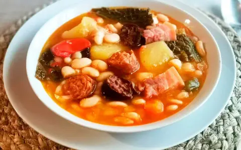 Un reconfortante cocido montañés, rebosante de alubias blancas, patatas, chorizo, morcilla y tocino, servido en un cuenco blanco.
