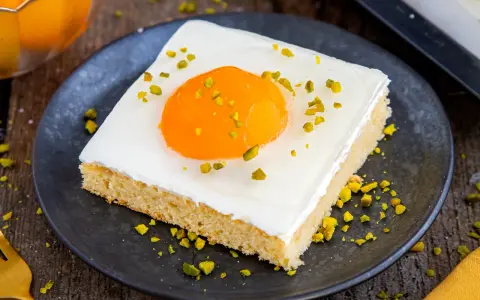 Ein Stück Kuchen mit weißer Glasur, einem orangen Gelee-Dotter und gehackten Pistazien, das wie ein Spiegeleikuchen mit Quark aussieht.