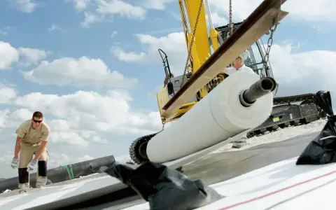 Excavadora descarga un rollo de lámina bentonita. Obreros trabajan en la instalación bajo un cielo nublado.