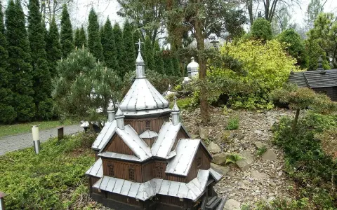 Drewniana cerkiewka z metalowym dachem, miniatura w parku miniatur Myczkowce, otoczona zielenią i kamieniami.
