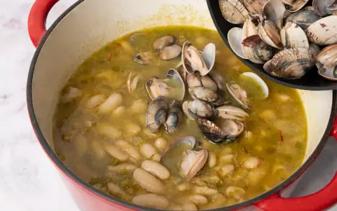 Alubias cremosas con almejas en una olla roja, añadiendo más almejas de una sartén.