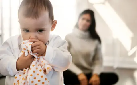 Kleines Baby hält sich an einem Tuch fest, wirkt schüchtern. Im Hintergrund sitzt eine unscharfe Frau.