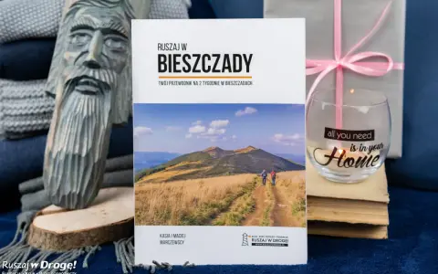 Przewodnik "Ruszaj w Bieszczady" z widokiem na góry i turystów. Obok rzeźba z drewna i świeca z napisem "all you need is in your Home".