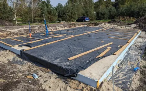Gotowy fundament pod dom szkieletowy, z czarną folią i drewnianymi belkami, czeka na dalsze prace budowlane.