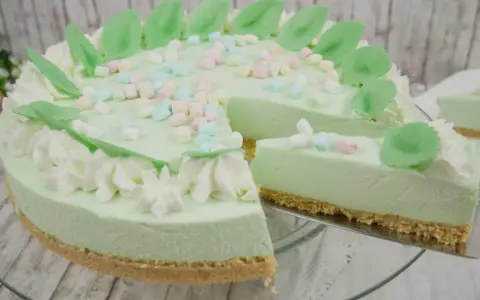 Eine helle, cremige Philadelphia Torte Waldmeister mit bunten Marshmallows und grünen Blättern verziert. Ein Stück wird gerade herausgeschnitten.