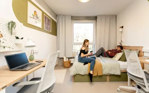 Dwie osoby w pokoju studenckim, jeden leży na łóżku, drugi siedzi obok. Tak wyglądają egzaminy na studiach – chwila wytchnienia przed nauką.