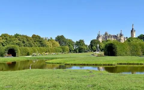 Ein Reiher steht am Ufer eines Teichs im Schweriner Schlossgarten. Im Hintergrund thront das Schloss über grünen Bäumen.