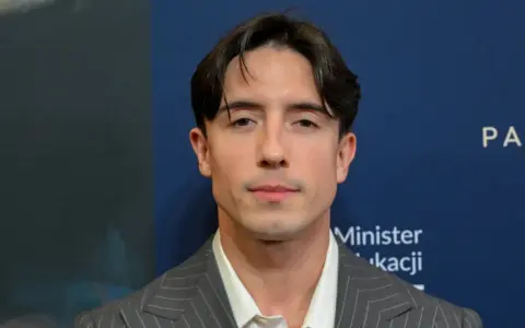 Dawid Kwiatkowski w szarym garniturze na tle niebieskiej ściany z napisami.