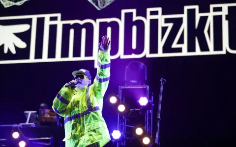 Fred Durst z Limp Bizkit na koncercie, w neonowej kurtce, z ręką w górze. Tło to logo zespołu.