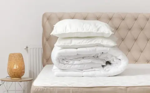 Edredón blanco doblado con almohadas encima, listo para lavar edredón en lavadora. Cabecero acolchado y lámpara de mimbre.