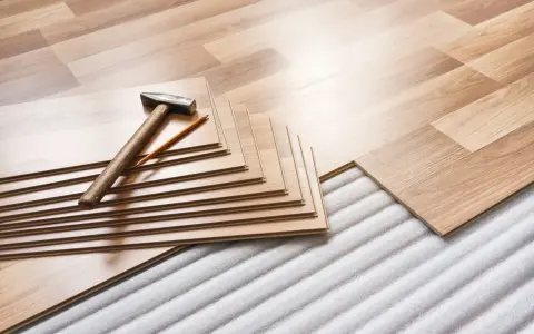 Tablas de suelo laminado apiladas con martillo y lápiz, listas para aislar suelo frío sobre una base acolchada.