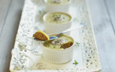 Trois ramequins de foie de volaille, ornés de champignons et de persil, sur un plateau ajouré. Une cuillère présente une bouchée de ce délice.