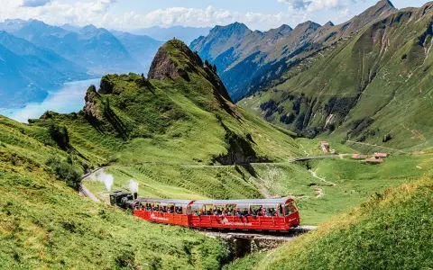 A red Brienz Switzerland train winds through lush green mountains, with a stunning blue lake in the distance.