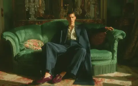 Giovane uomo con abito blu e camicia fantasia, seduto su un divano verde smeraldo, indossa un elegante abito con mocassino bordeaux.