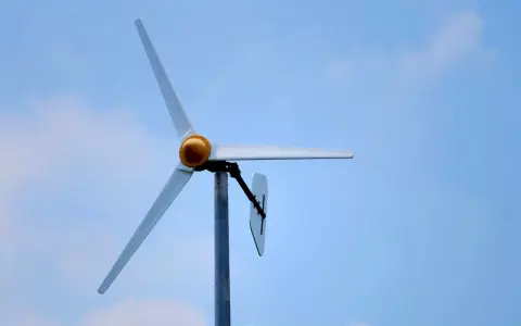 Biała elektrownia wiatrowa z żółtym elementem na środku, na tle błękitnego nieba z chmurami.