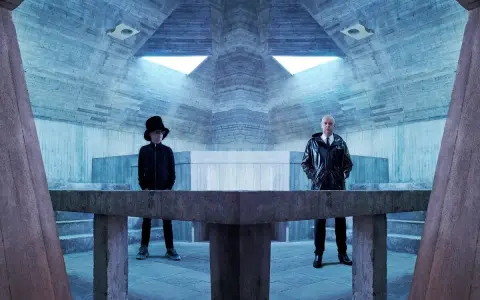 Pet Shop Boys in futuristischer Betonarchitektur. Neil Tennant mit Zylinder, Chris Lowe im glänzenden Mantel.