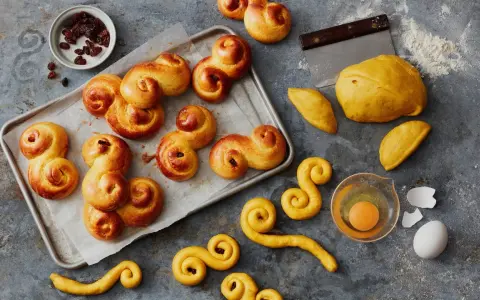 En bricka med gyllene, nybakade lussekatterna, redo att avnjutas. Bredvid ligger ingredienser som russin, deg och ett ägg. Dessa är de godaste lussekatterna!