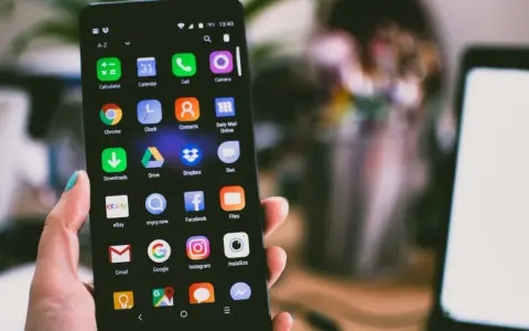 Dłoń trzyma smartfon z widocznym ekranem pełnym ikon aplikacji, w tym ikoną wiadomości SMS.