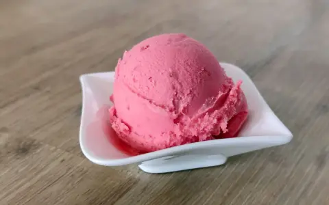 Ein Löffel cremiges Himbeersorbet in einer weißen Schale auf Holzhintergrund.