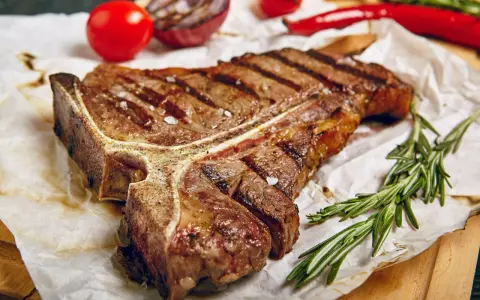 Saftiges T-Bone-Steak, perfekt gegrillt, mit grobem Salz bestreut, daneben Rosmarin, Tomaten und Chili.