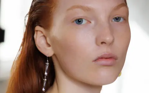 Primo piano di una giovane donna con capelli rossi e occhi azzurri, un neo della bellezza sul viso. Indossa un orecchino pendente.