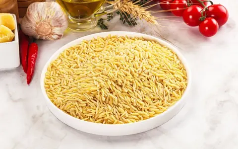 Miseczka pełna makaronu orzo, idealnego do przygotowania pysznego dania. Obok czosnek, papryczki chili, pomidorki i oliwa.