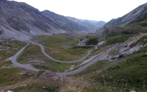 Strada tortuosa che sale verso il colle dei morti, tra montagne verdi e rocciose. Due ciclisti affrontano le curve.