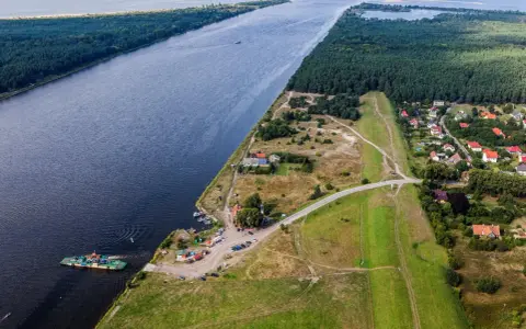 Widok na Wisłę i jej ujście. Prom przewozi samochody, a w oddali widać las i zabudowania.