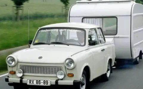 Biały Trabant ciągnie przyczepę kempingową po drodze.