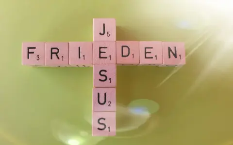 Buchstabensteine bilden ein Kreuz mit den Wörtern "FRIEDEN" und "JESUS". Ein Zitat aus Johannes 14.