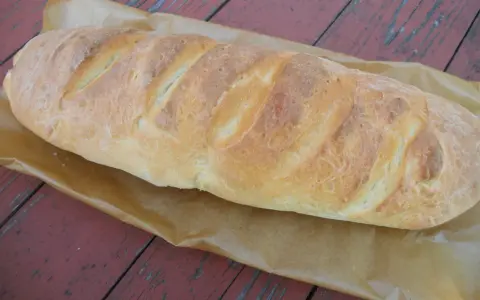 Złocista bułka paryska, świeżo upieczona, leży na papierze do pieczenia na drewnianym stole.