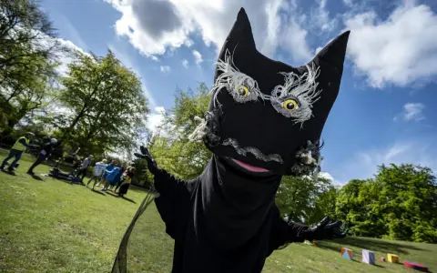 En stor, svart docka som liknar Syrsommarskuggan vinkar glatt i en park med folk i bakgrunden.