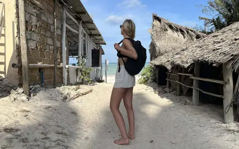 Kobieta z plecakiem na piaszczystej plaży, zastanawia się co zabrać na Zanzibar. W tle chatki z palmowymi dachami i budynek z kamienia.