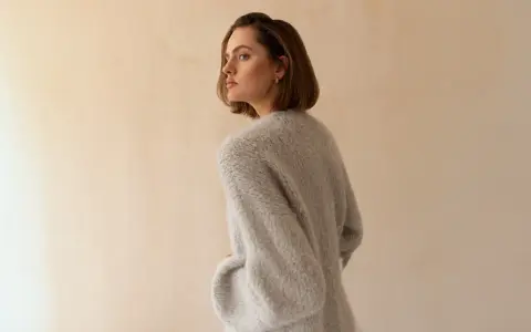 Młoda kobieta w szarym, wełnianym swetrze, który wygląda, jakby był ręcznie robiony. Delikatne światło podkreśla fakturę dzianiny.