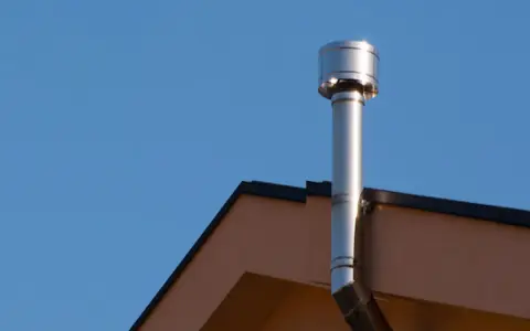 Scarico scaldabagno a gas in facciata, un tubo metallico lucido che sale verso il cielo azzurro, fissato al muro di un edificio.