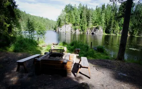 En mysig eldstad med bänkar vid en stilla sjö, omgiven av grönskande skog. Ett perfekt utflyktsmål i Dalarna för avkoppling.