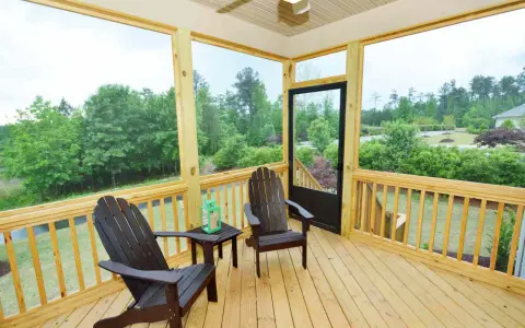 A screened-in porch with two Adirondack chairs, a small table, and a green lantern. The porch offers a view of lush trees and a suburban landscape.