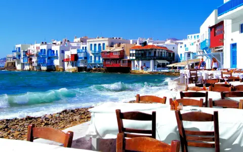 Cosa vedere a Mykonos in crociera: case colorate sul mare, onde che si infrangono sulla spiaggia di ciottoli e tavoli apparecchiati per godersi il panorama.