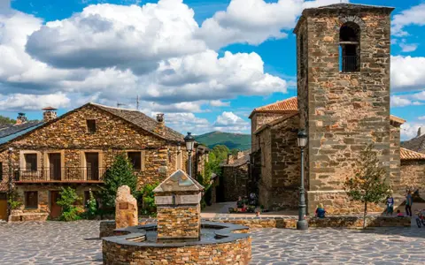 Plaza con fuente de piedra y campanario en un pueblo de la ruta negra Guadalajara. Casas de piedra y cielo azul con nubes.