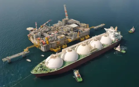 Praca na statku LNG: potężna jednostka z kopułami ładunkowymi przy platformie wydobywczej, holowniki asystują.